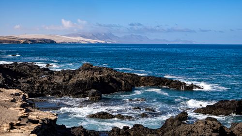 Free A stunning view of a rocky coastline with waves crashing against the shore under a clear blue sky. Stock Photo
