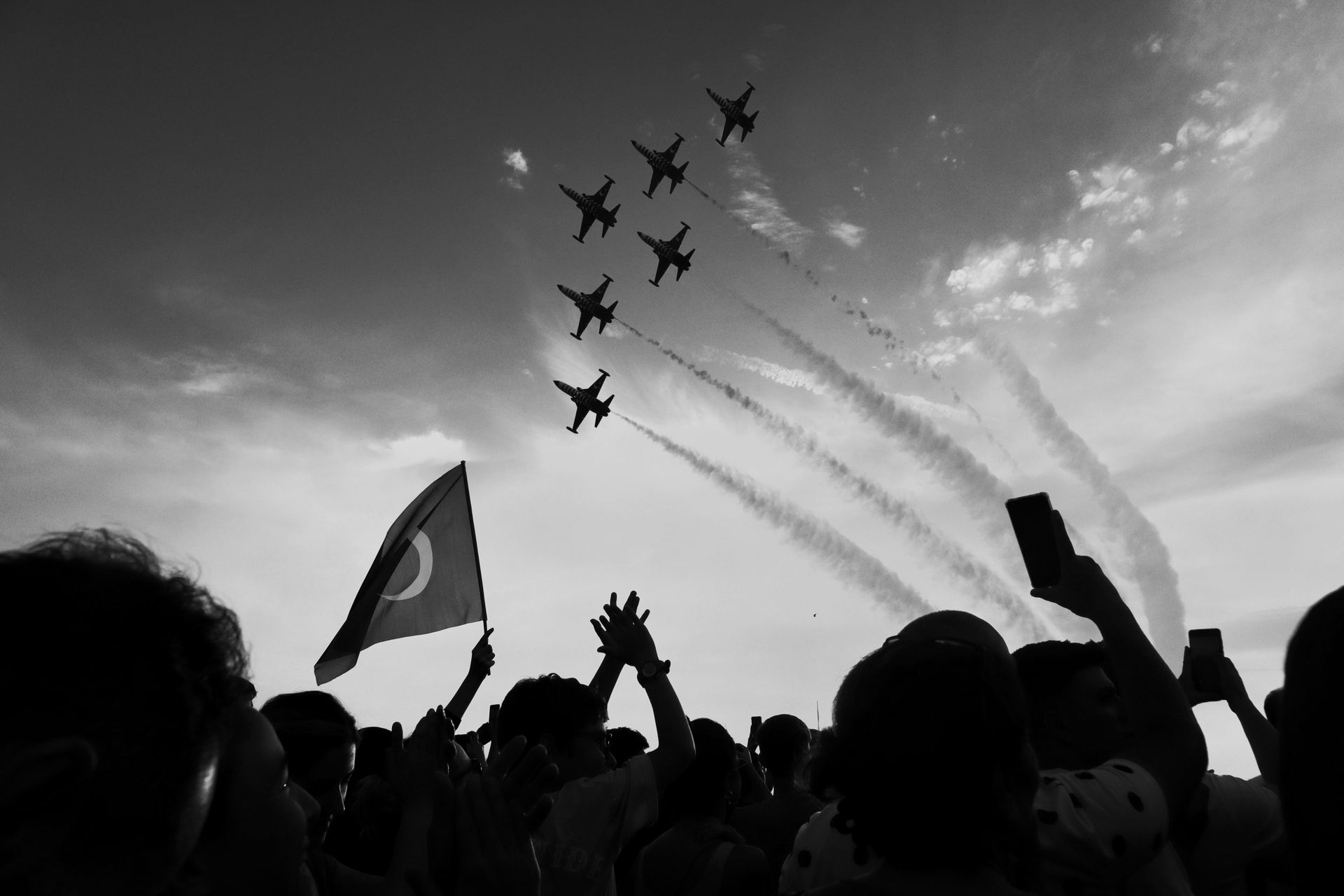 Silhouette of jets performing aerobatics over a cheering crowd with a Turkish flag.