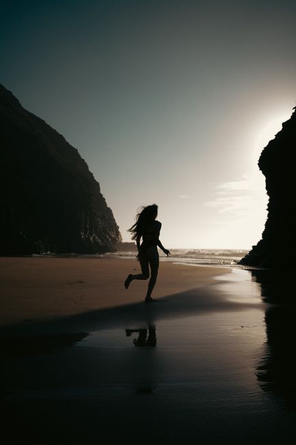 A woman runs along a secluded beach with ocean cliffs at twilight, creating a stunning silhouette.