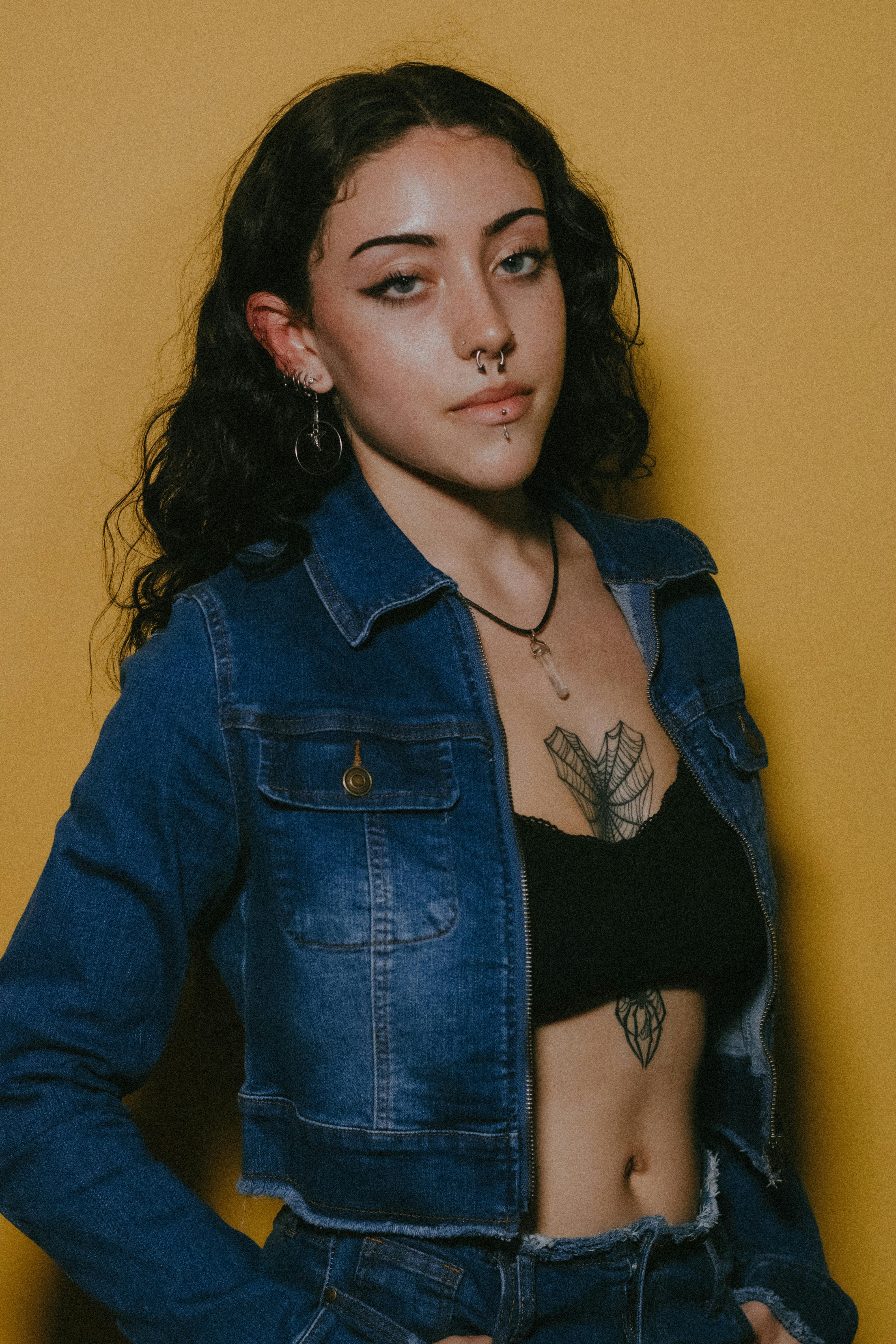 Young woman with tattoos posing in denim jacket against a vibrant yellow background.