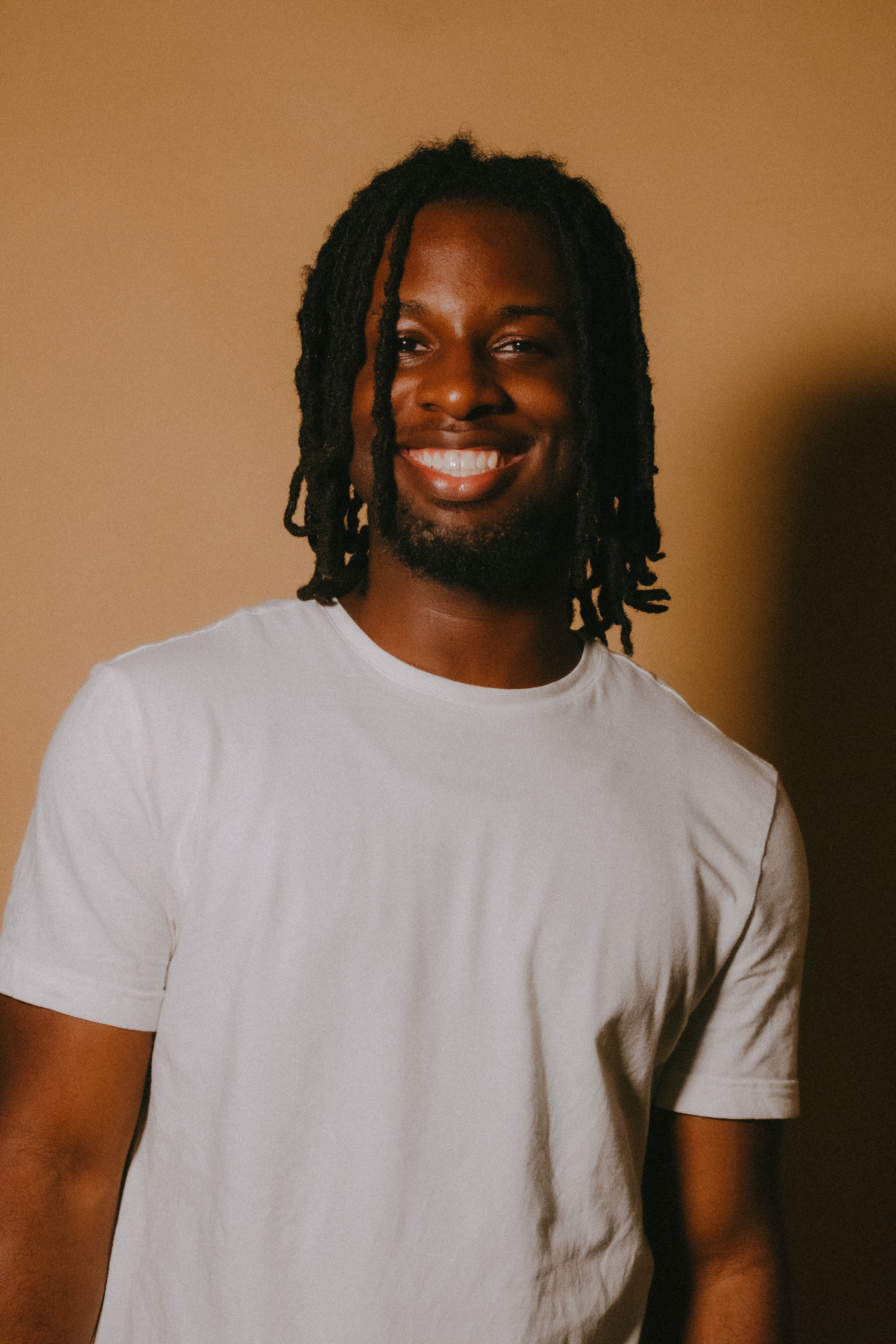 Smiling man with dreadlocks in a studio setting wearing a white shirt.