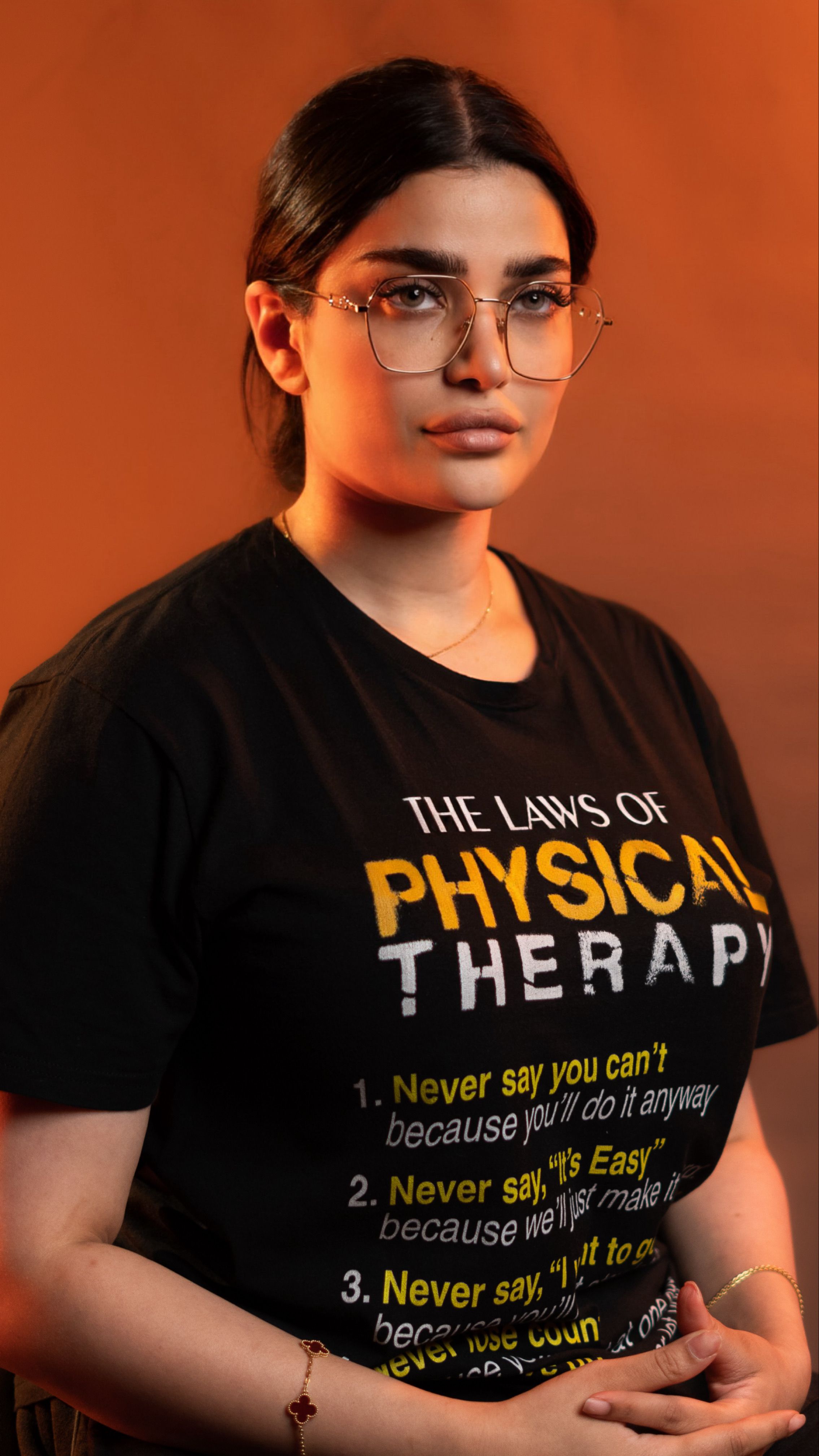 Confident woman in glasses wearing a 'The Laws of Physical Therapy' t-shirt on a warm background.