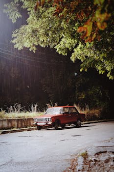 A classic vintage car parked under a tree on a rainy street at night, with an atmospheric and moody feel.