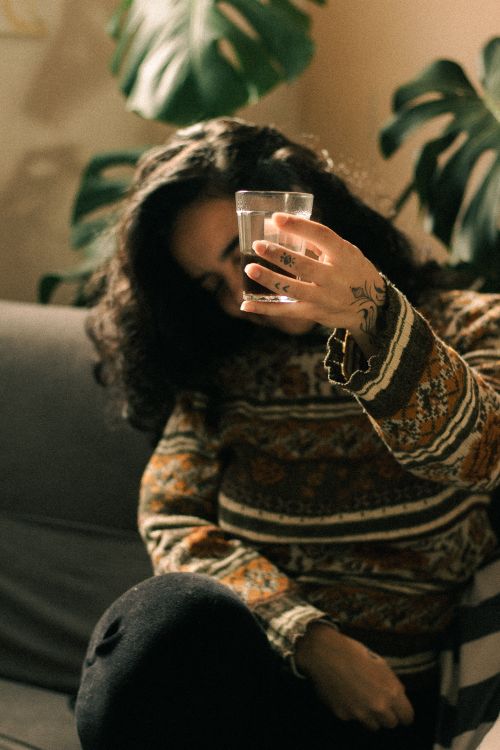 Free A woman in a cozy sweater holding a glass, framed by leafy plants indoors. Stock Photo