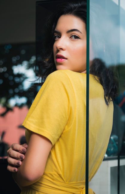 Elegant portrait of a woman in yellow posing by a glass pane, reflecting light.