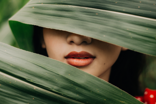 Close-up of a woman's lips hidden behind lush green leaves, evoking a sense of mystery and nature.