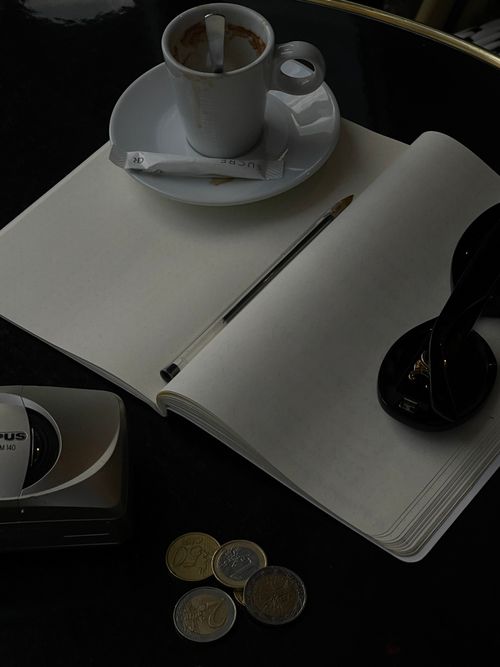Free An artistic shot of a café table featuring a cup of espresso, notepad, sunglasses, and coins. Stock Photo