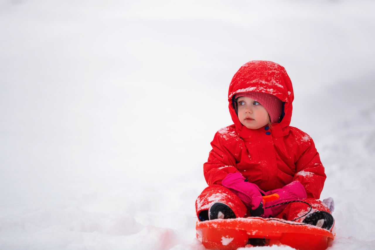 Toddler in a red one-piece snowsuit with mittens sitting on a sled in the snow