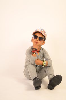 Confident young boy in a fashionable outfit posing in a studio setting with sunglasses and a cap.