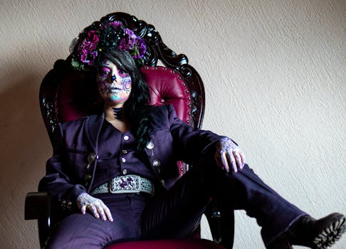 Woman in purple suit with Catrina makeup, seated in ornate chair, celebrating Dia de los Muertos.