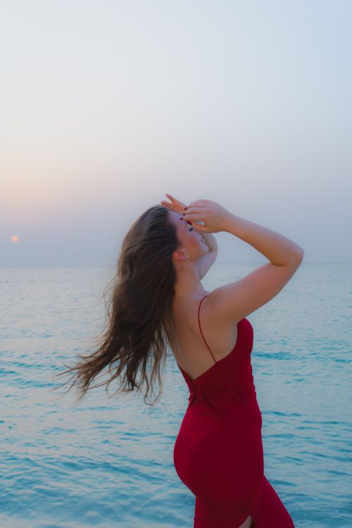 Free A woman in a red dress poses elegantly on the beach, with her hair blowing in the sea breeze at sunset. Stock Photo