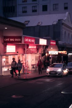 Urban night street view featuring people and illuminated Café BBQ Duck sign in a city.