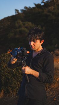 A man in a black shirt holding a camera outdoors during sunset.