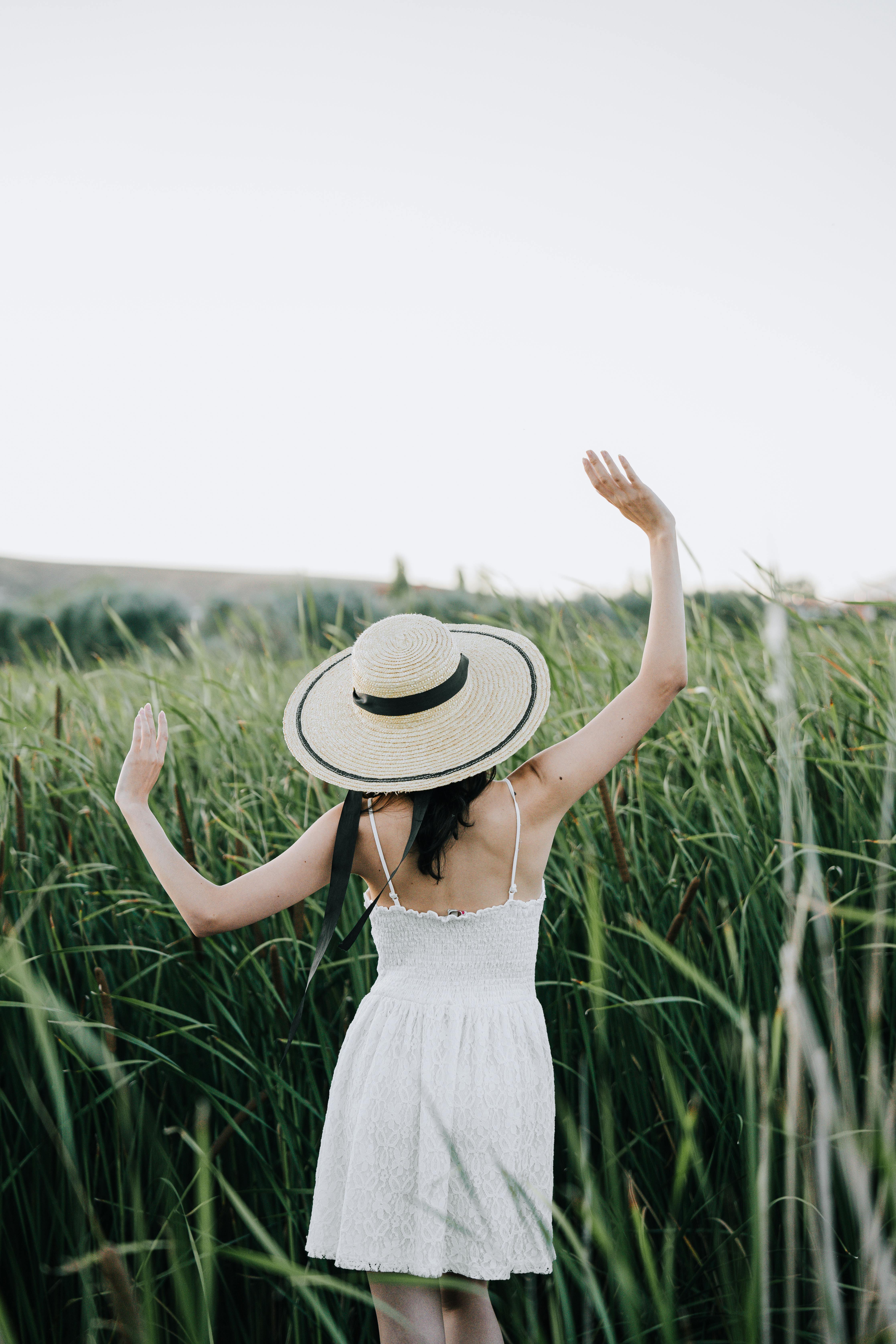 Back view of woman in hat and white dress standing in tall grass on a sunny day.
