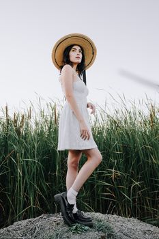 Stylish young woman in a white dress and sun hat stands in tall grass, embracing summer vibes.