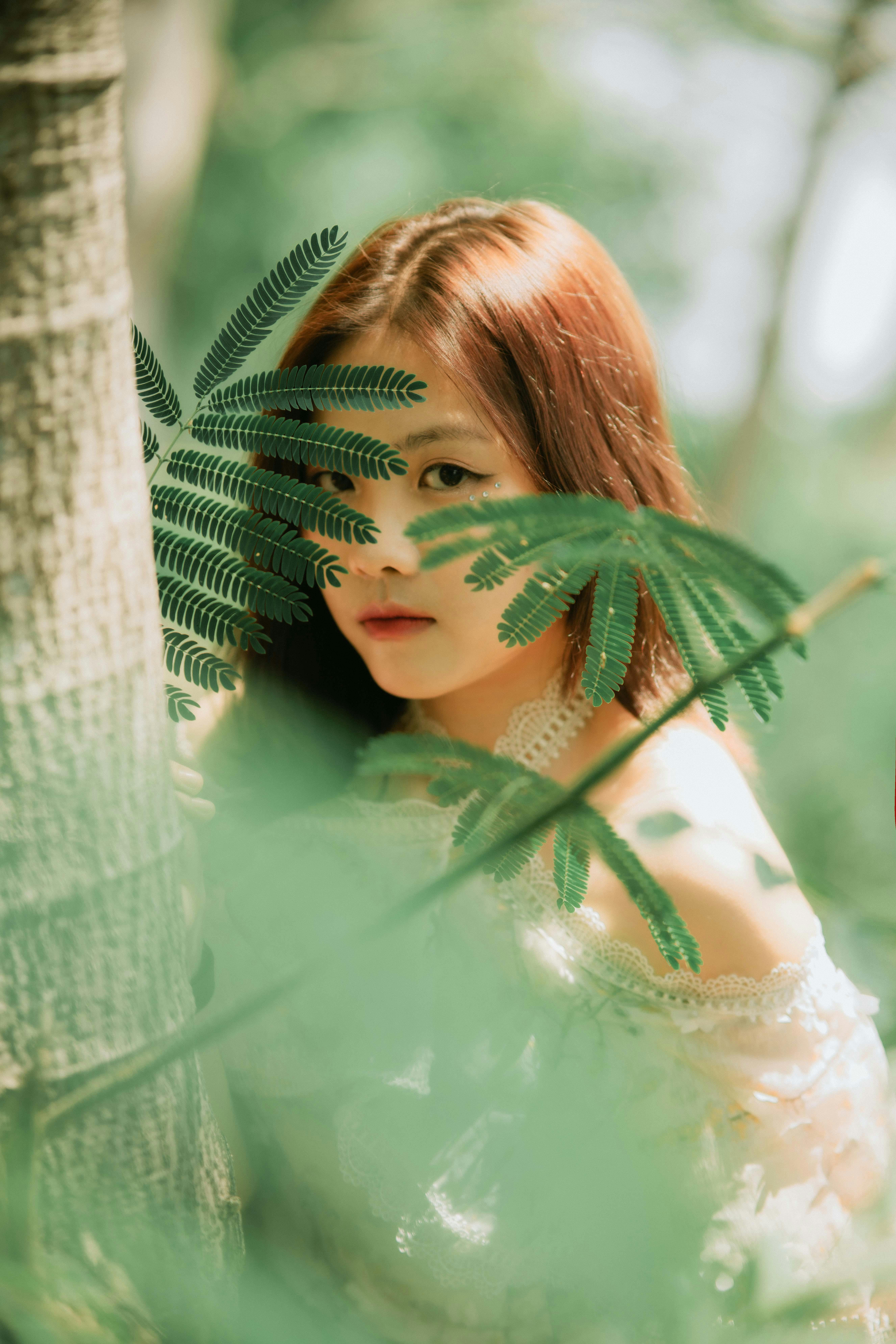 Photo of a young woman partially hidden by leaves, with summer sunlight creating a serene atmosphere.