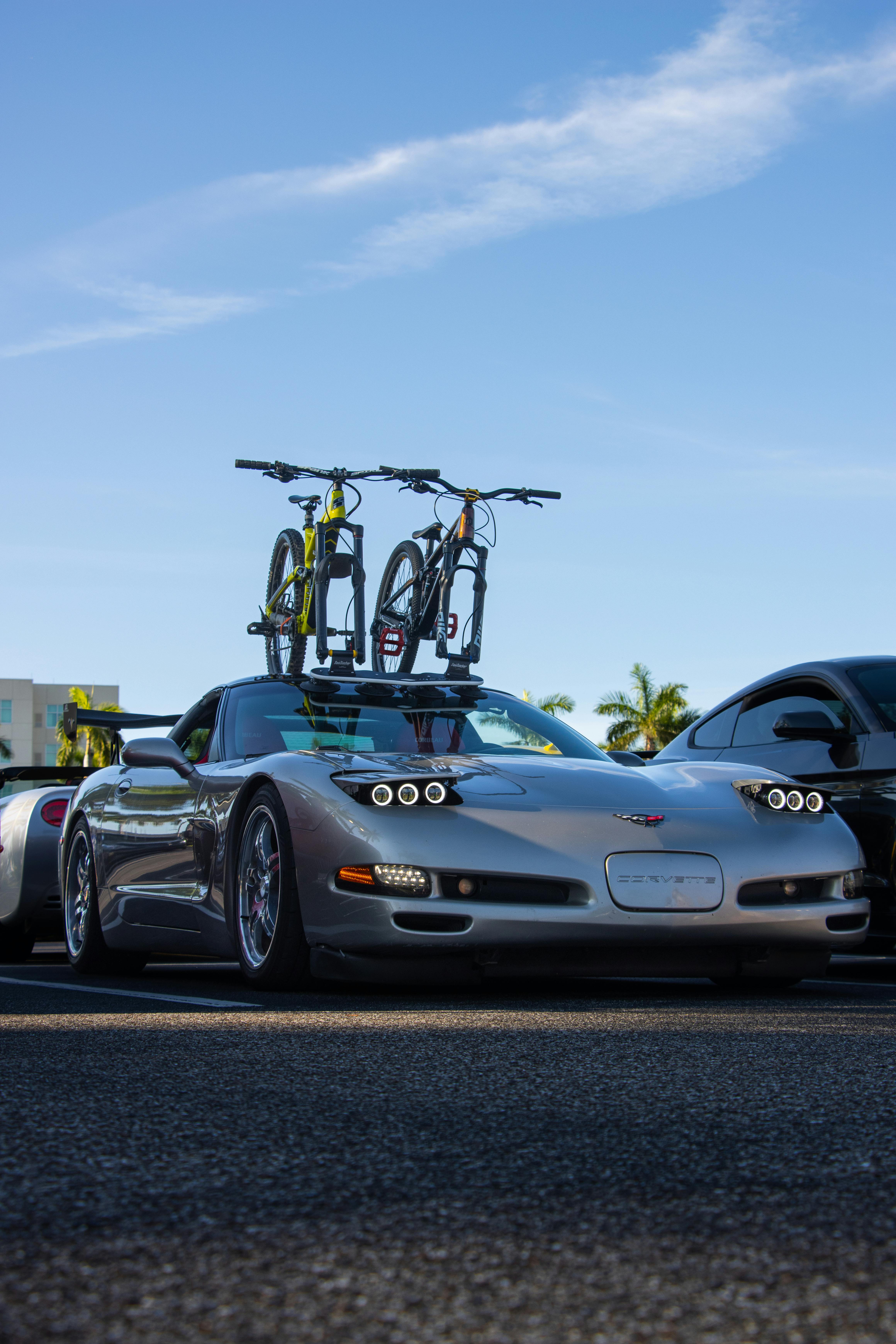 Silver sports car with bicycles mounted on roof, parked under a clear blue sky in an urban environment.
