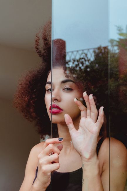 Portrait of a stylish woman with an afro and red lipstick, looking through a glass window, captured indoors.