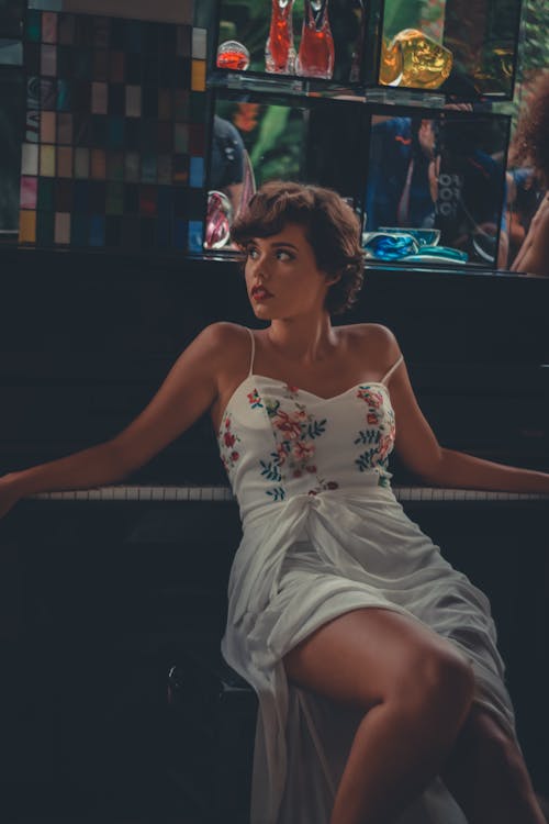 피아노 옆에 의자에 앉아 흰색과 여러 가지 빛깔의 꽃 스파게티 스트랩 드레스를 입고 여자