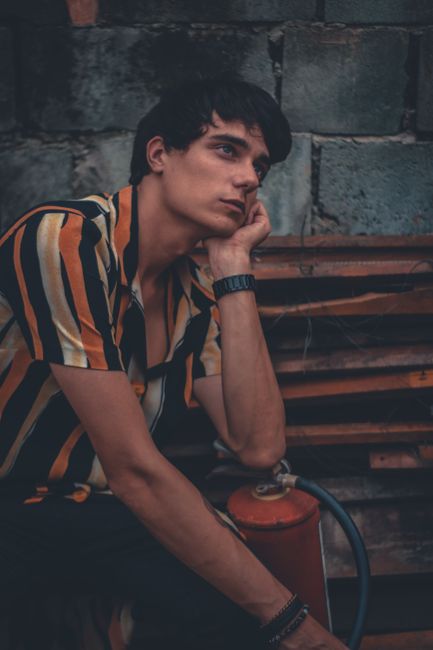 A young man in a striped shirt, deep in thought, sitting against a rustic background.