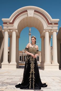 An elegant woman poses in a luxurious black gown with gold embellishments, framed by classical architecture.