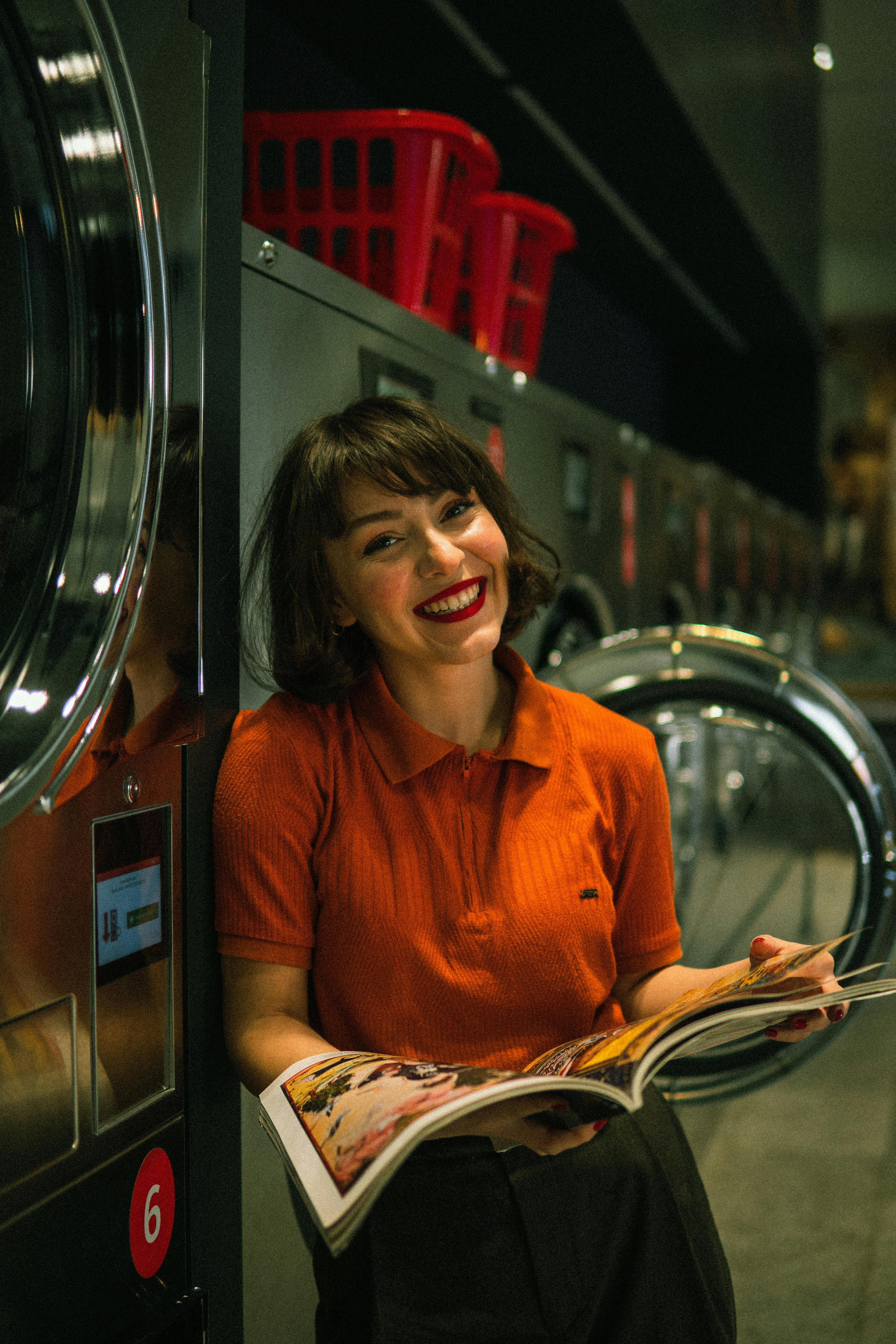A cheerful woman in an orange shirt enjoys a magazine while doing laundry.