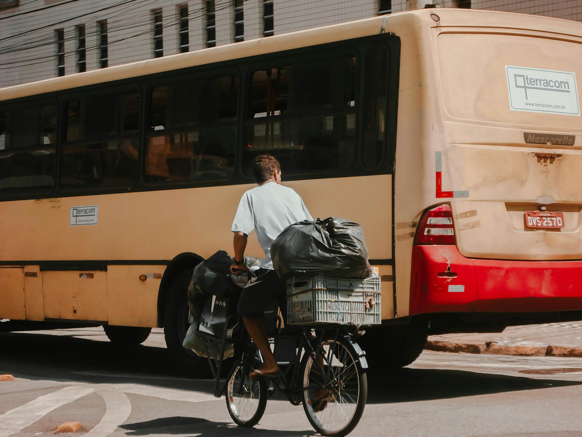 Man rides bicycle with cargo beside a bus in an urban street setting under bright sunlight.