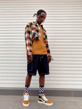 Fashionable young man with locs in plaid shirt and sneakers against a white garage door.