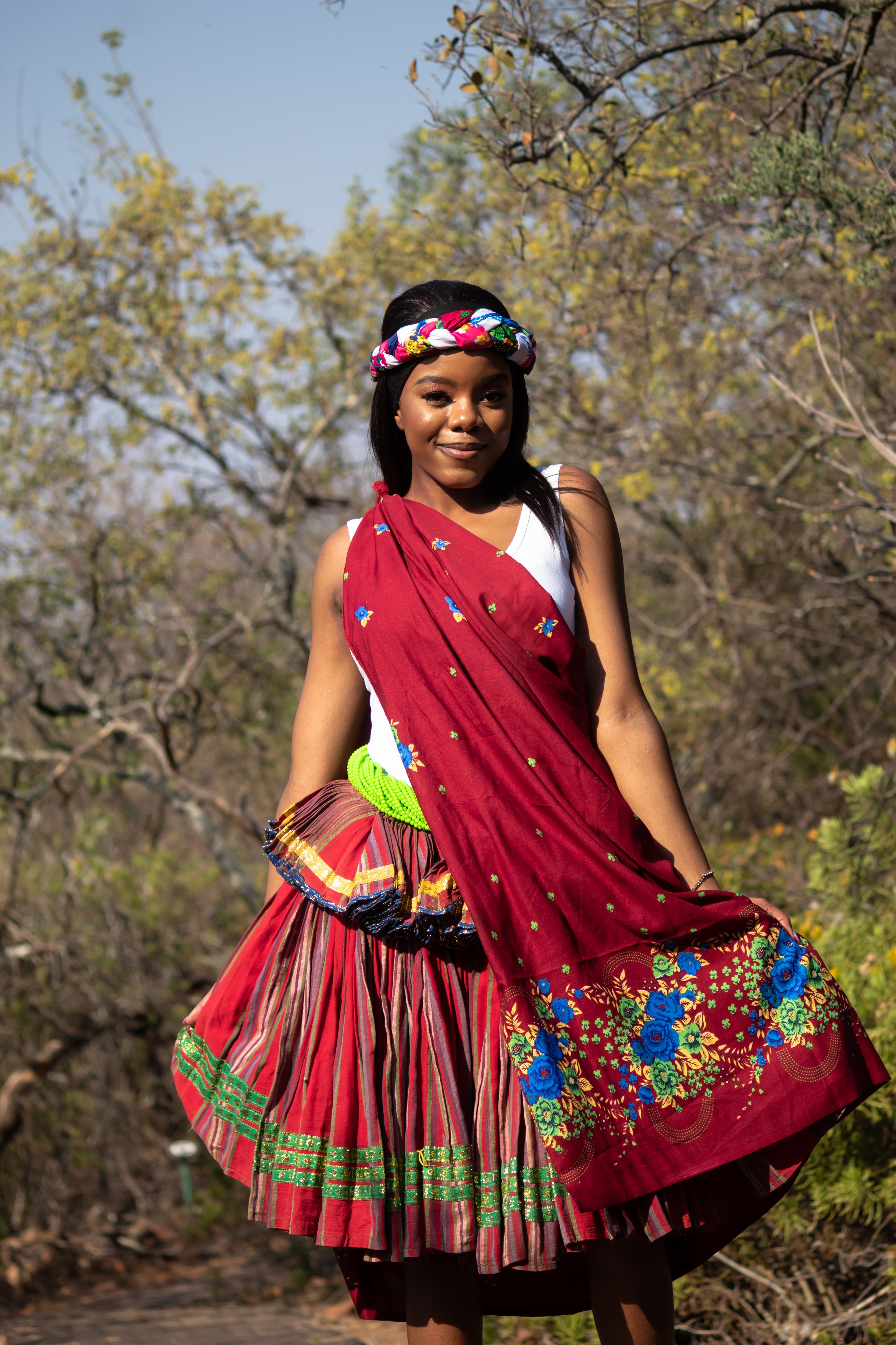 Smiling woman in vibrant traditional clothing standing in a sunlit forest, embracing cultural heritage.