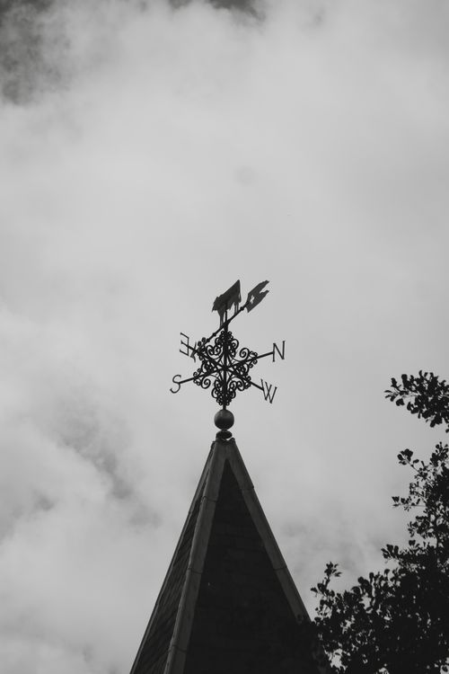 Free Black and white photo of a weather vane atop a gothic tower against a cloudy sky in London. Stock Photo