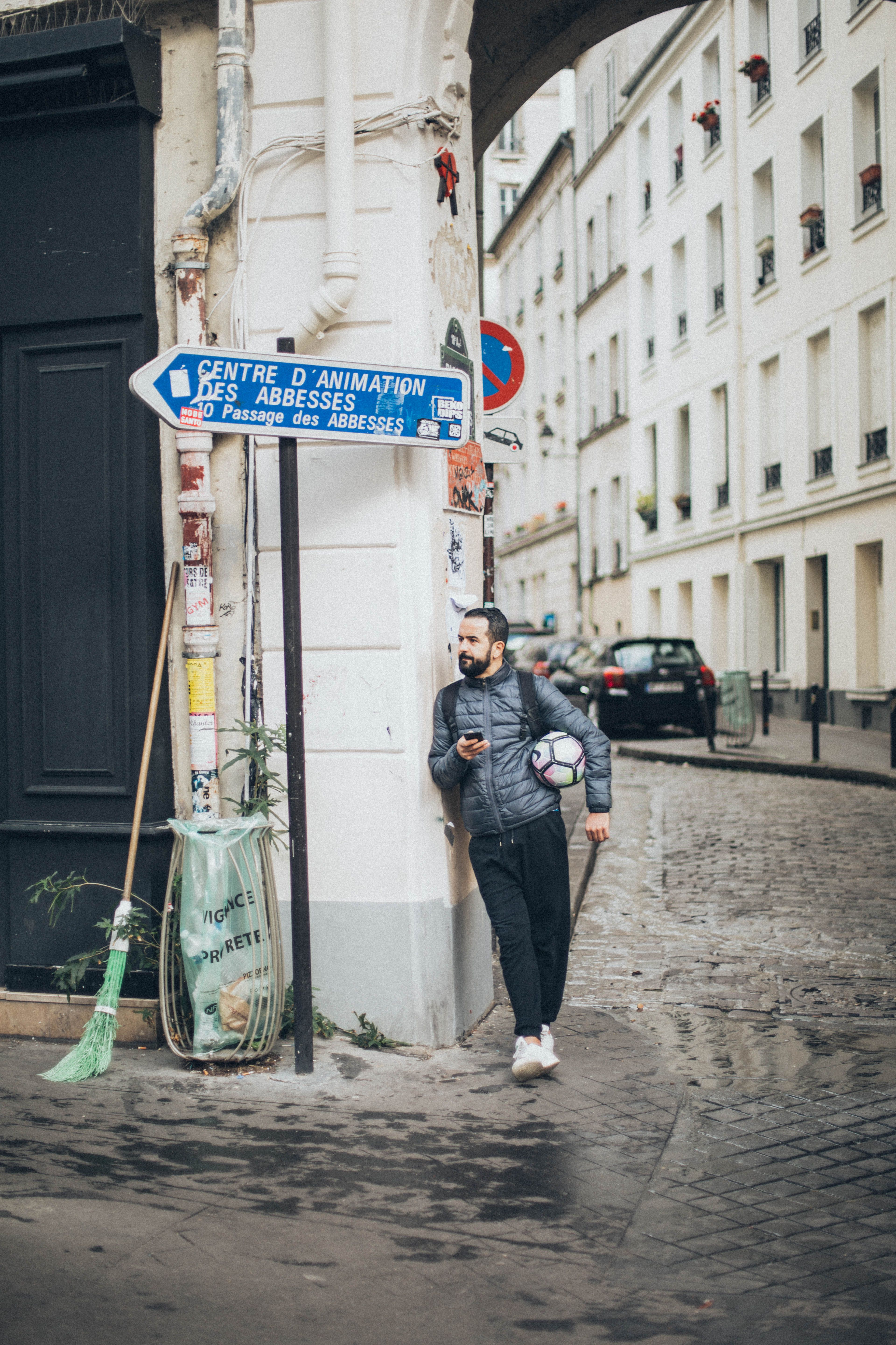 Man leaning against a wall on a cobblestone street holding a soccer ball and phone in a European city.