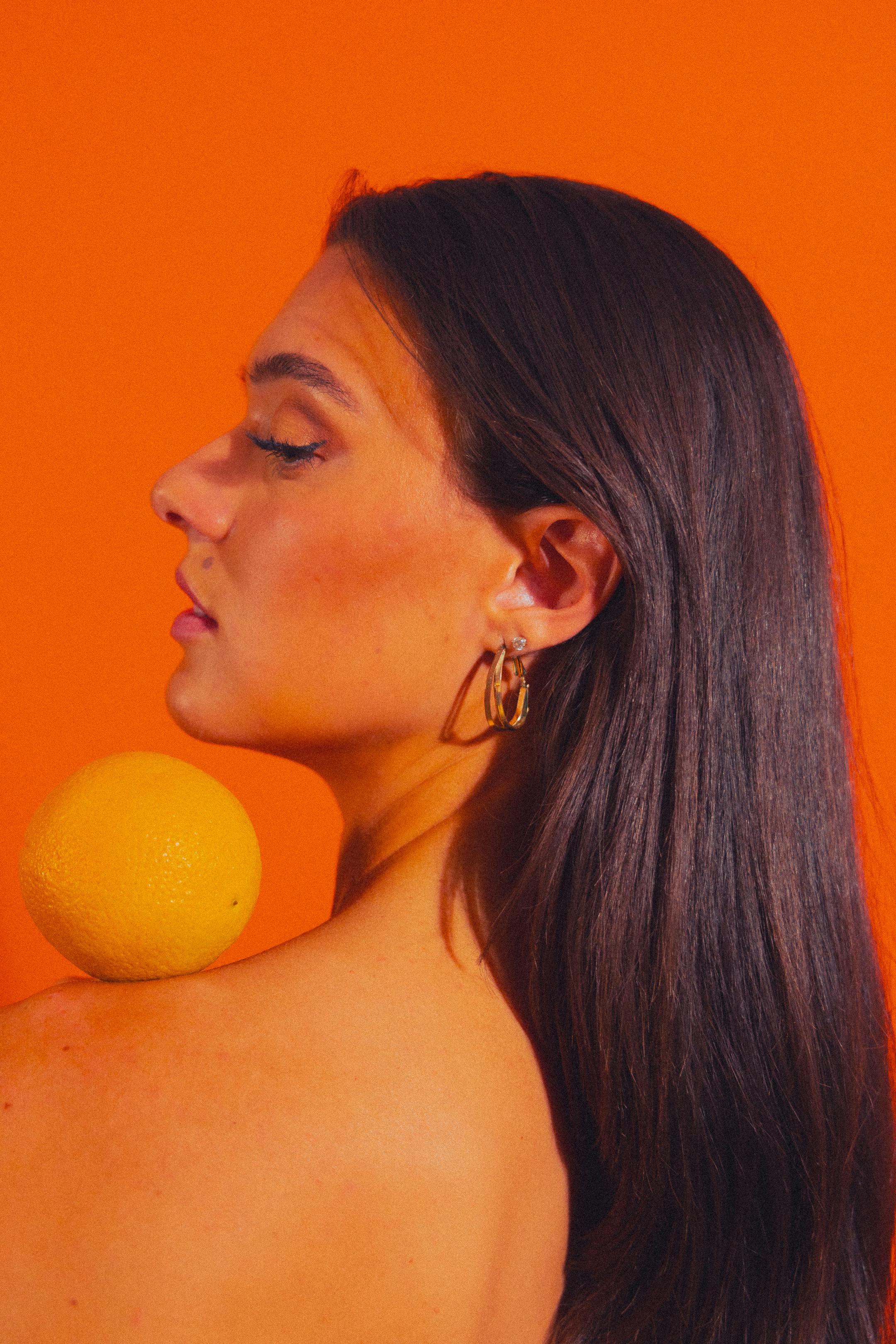 A striking portrait of a woman with long dark hair, styled against a bold orange background with a fruit accent.