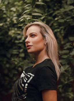 一位金发女子身着黑色衬衫，在郁郁葱葱的绿植环绕中，于户外优雅地摆出时尚肖像的姿势