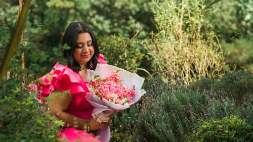 Free A woman in a bright pink dress holding a bouquet of flowers, surrounded by lush green foliage. Stock Photo
