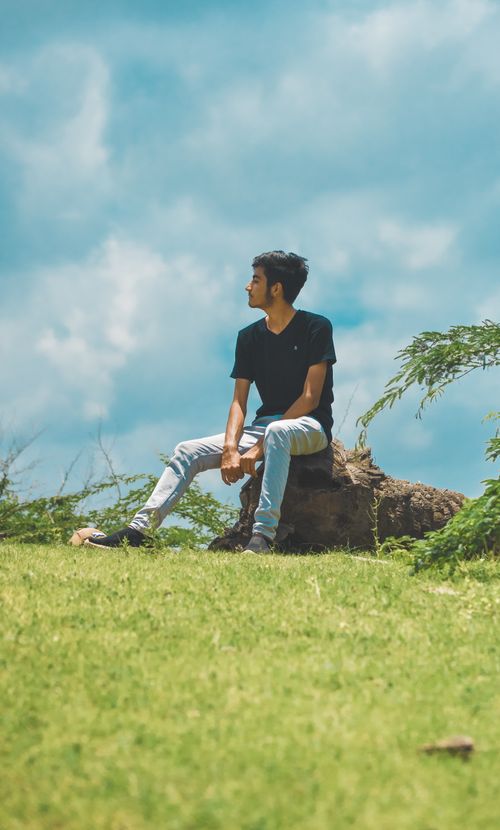 Free A young man sitting on a log outdoors, enjoying a sunny summer afternoon with a clear blue sky. Stock Photo