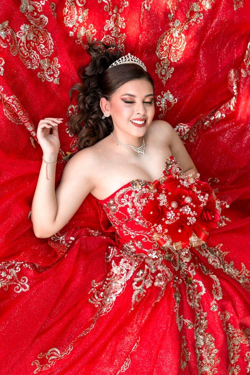 Free Portrait of a smiling woman in a stunning red gown adorned with a tiara and floral bouquet. Stock Photo