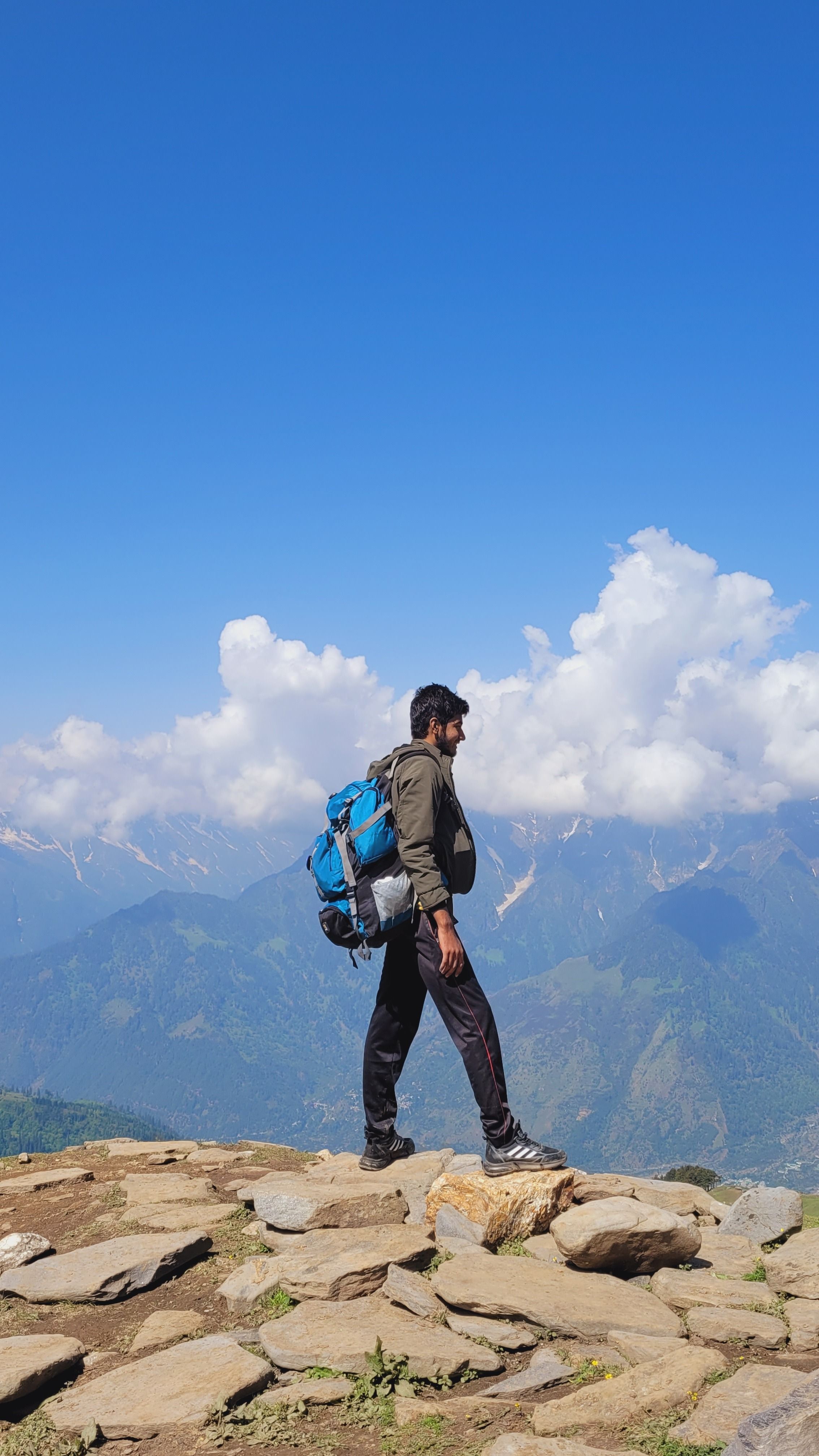 A hiker stands on a mountain summit with a blue backpack, surrounded by clouds and blue sky.