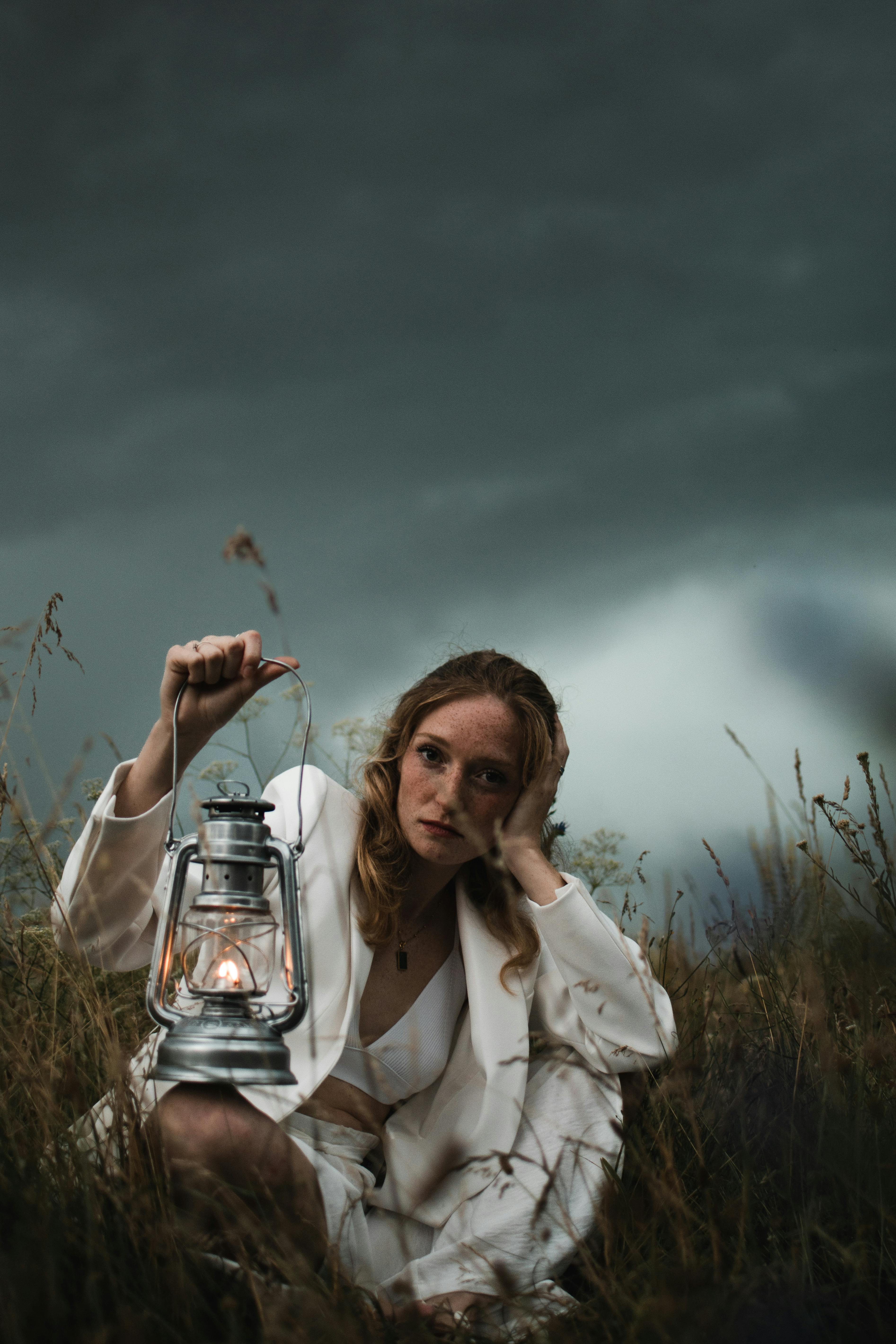Young woman in elegant white jacket holding a lantern in a field under stormy skies.