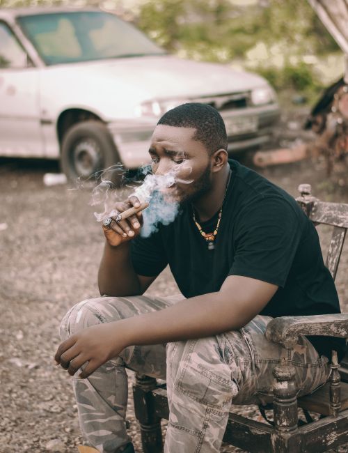 Free A man sitting outdoors, smoking a cigar in a rustic setting, with a car in the background. Stock Photo
