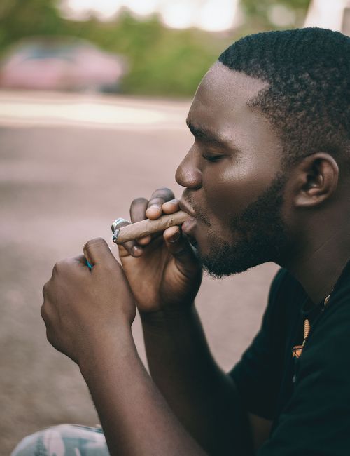 Free A young adult man with a beard smokes a cigar outdoors. Thoughtful expression. Stock Photo