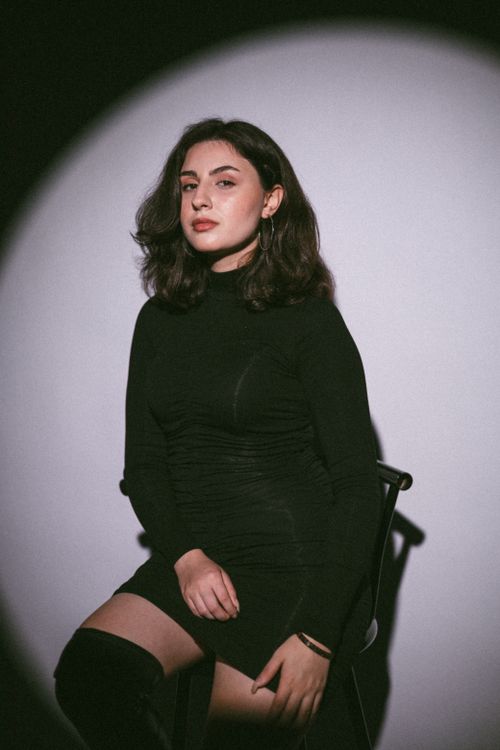 Free Stylish studio portrait of a woman in a black dress seated under spotlight, exuding elegance. Stock Photo