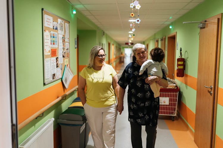  Elderly Woman With A Doll Led By Hand By A Caregiver Through The Nursing Home Corridor