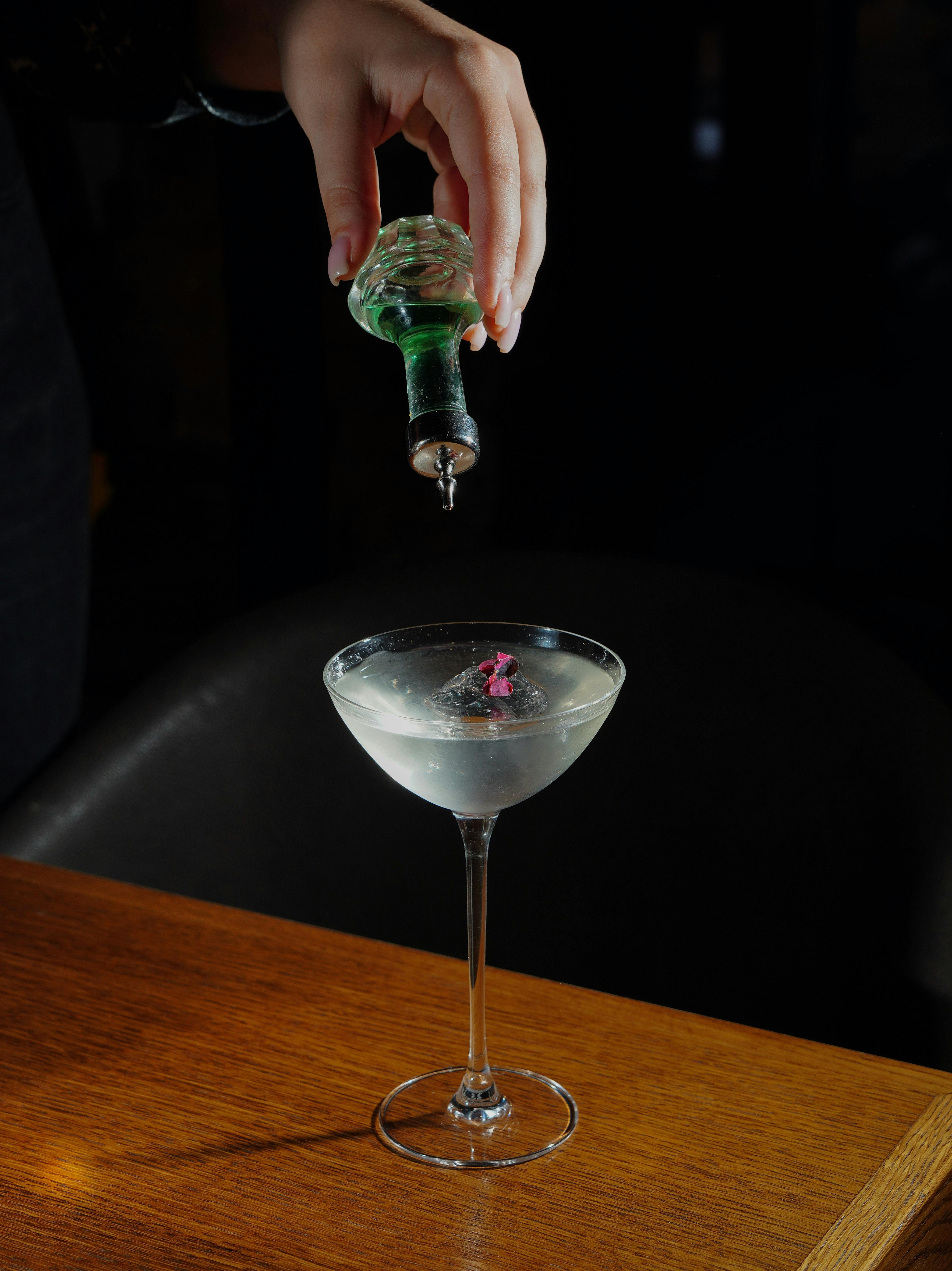 A close-up of a hand pouring gin from a bottle into a martini glass on a dark bar top.