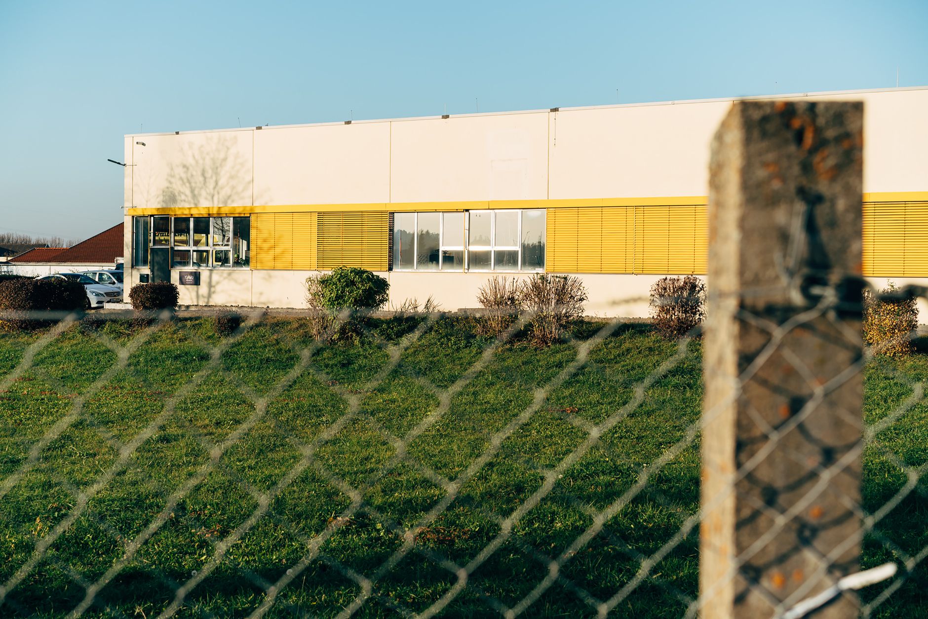 A modern industrial building with yellow accents and a chain link fence in the foreground.