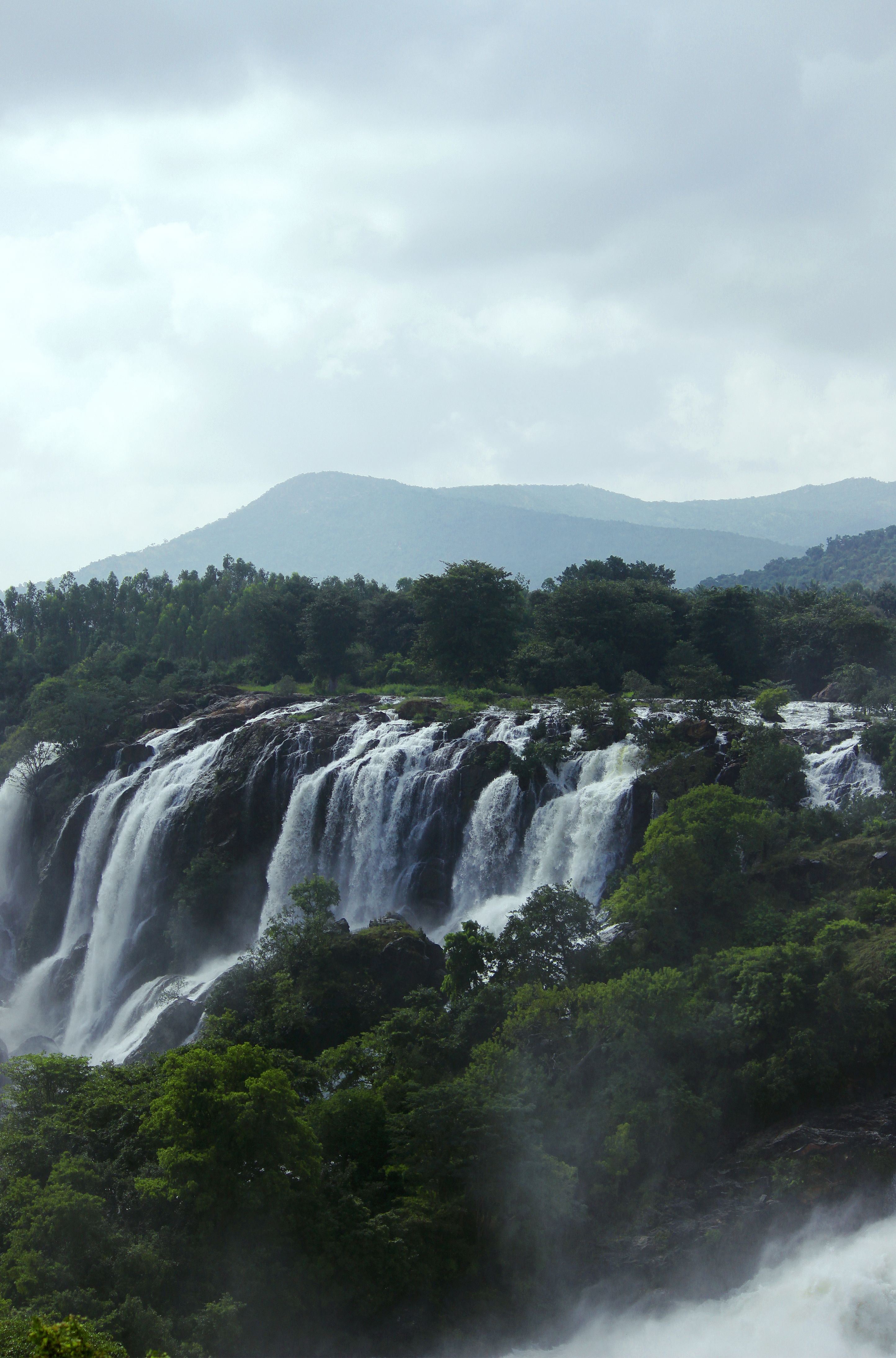 A breathtaking view of a waterfall flowing through dense green forest with mountains in the background.
