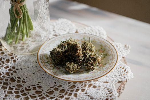 Dried cannabis buds on a vintage plate on a doily-covered surface, well-lit indoors.