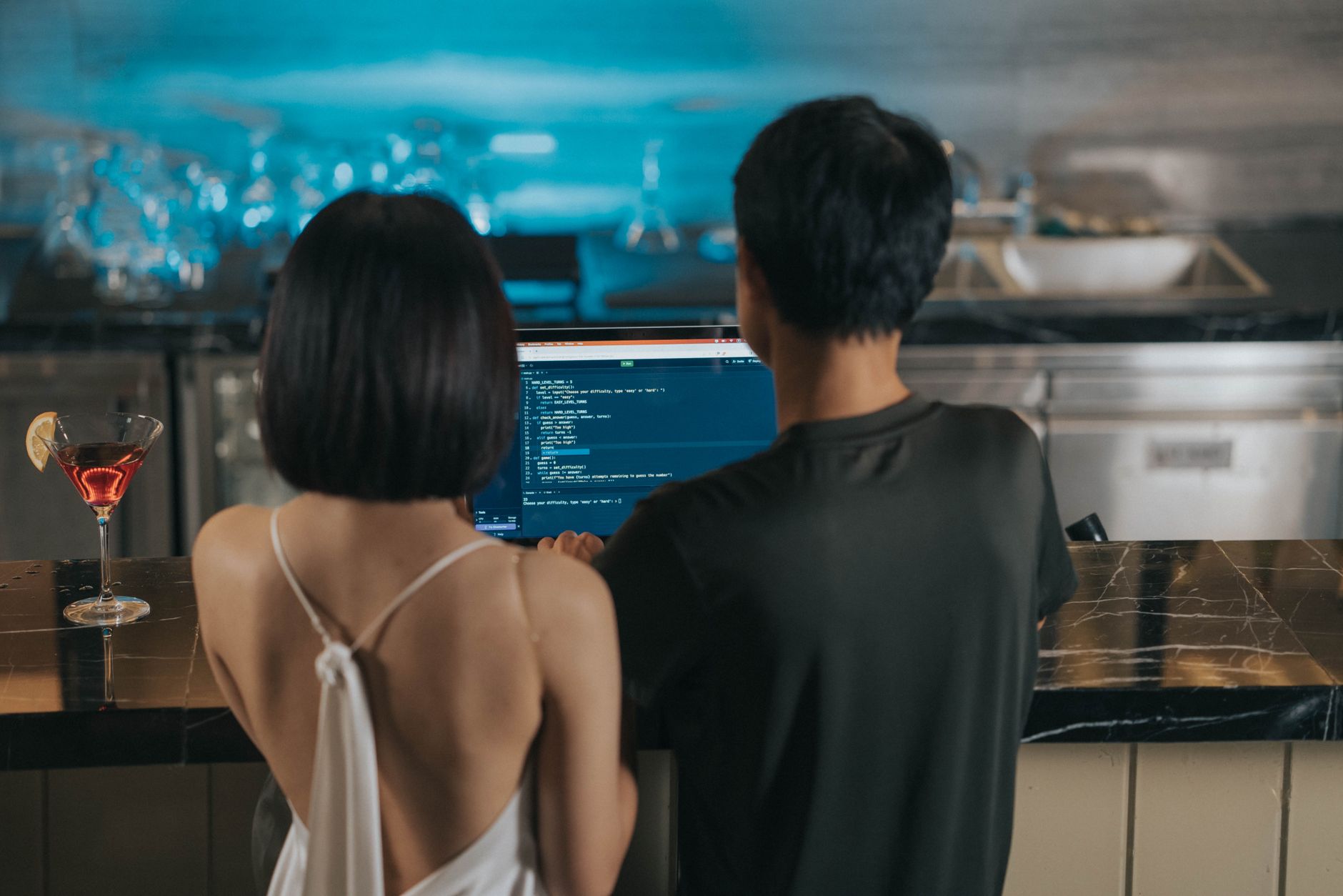 Couple coding together at a bar counter, drinks nearby, casual atmosphere.