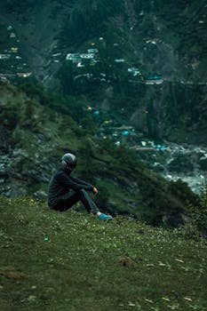 A lone figure sits on a green hilltop, gazing over a lush mountainous valley.
