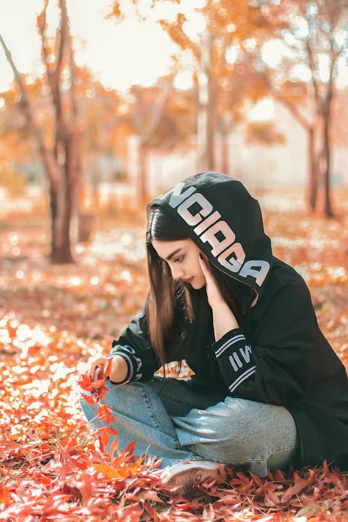 Girl Wearing a Hood Sitting on Ground with Red Autumn Leaves