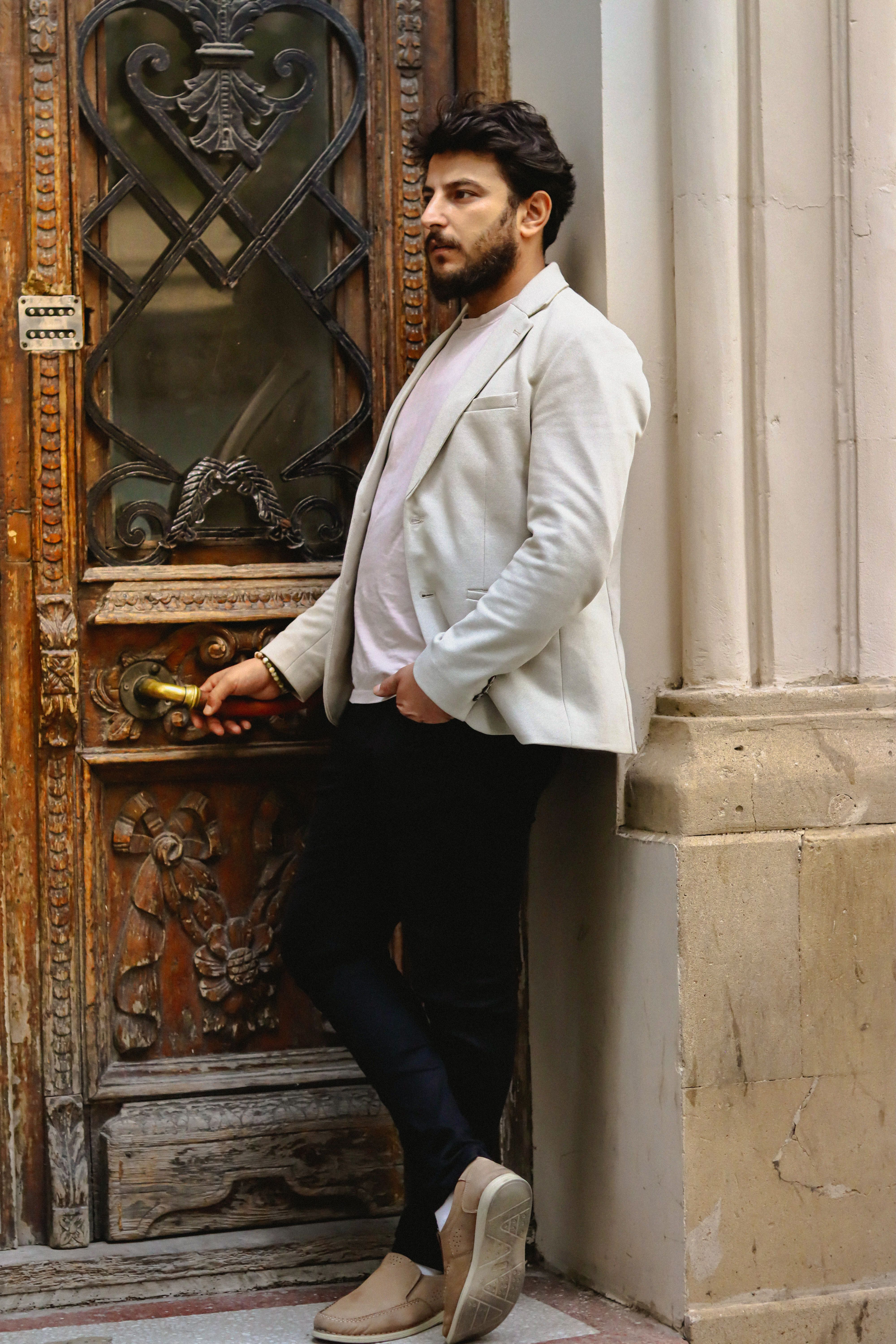 Stylish man in a white jacket poses by an ornate vintage door in an urban setting.